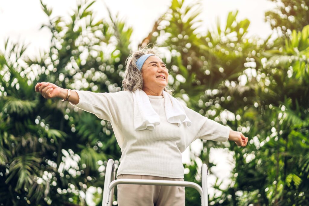 An elderly woman stands with arms outstretched, smiling joyfully in a vibrant green outdoor setting.