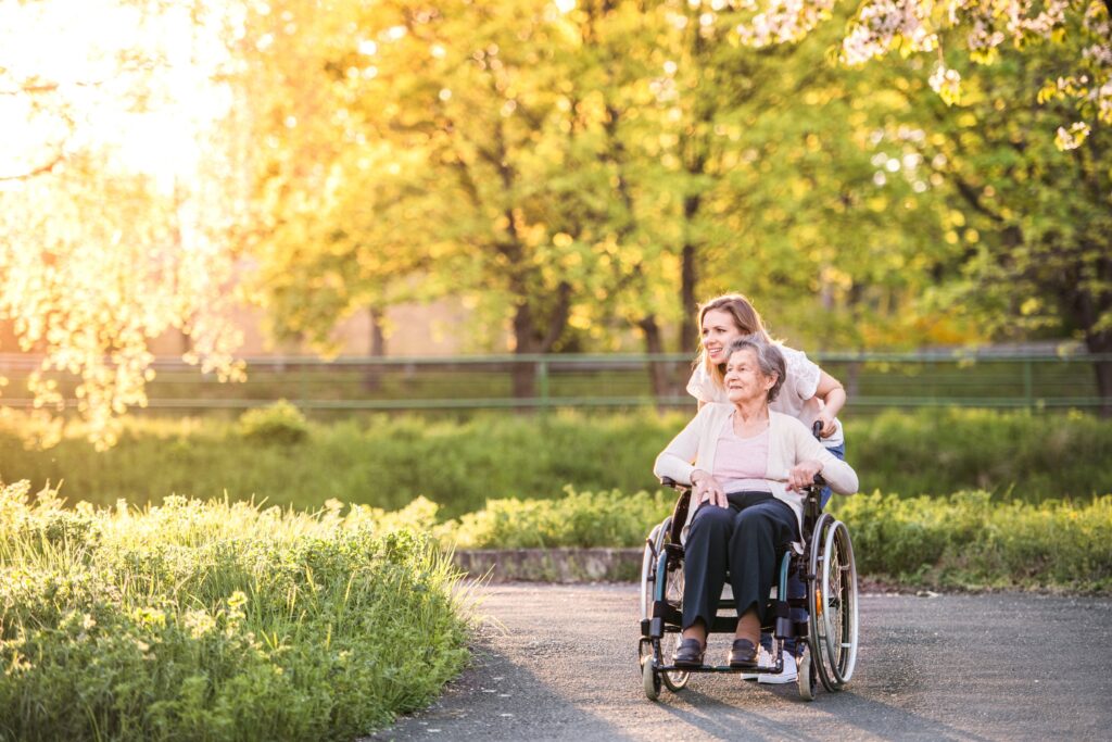 A young woman pushes an elderly woman in a wheelchair along a sunlit path surrounded by lush greenery.