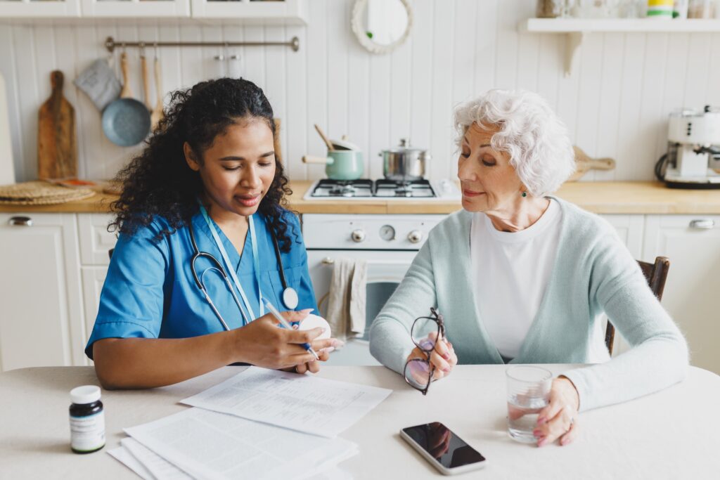 A healthcare professional discusses medication with an elderly patient at a kitchen table, emphasizing care and communication.