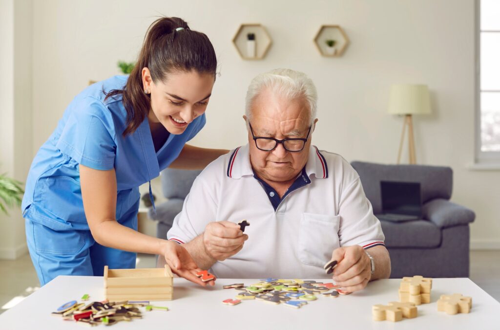 A caregiver assists an elderly man with a puzzle, fostering a warm interaction in a bright, cozy room filled with colorful pieces.